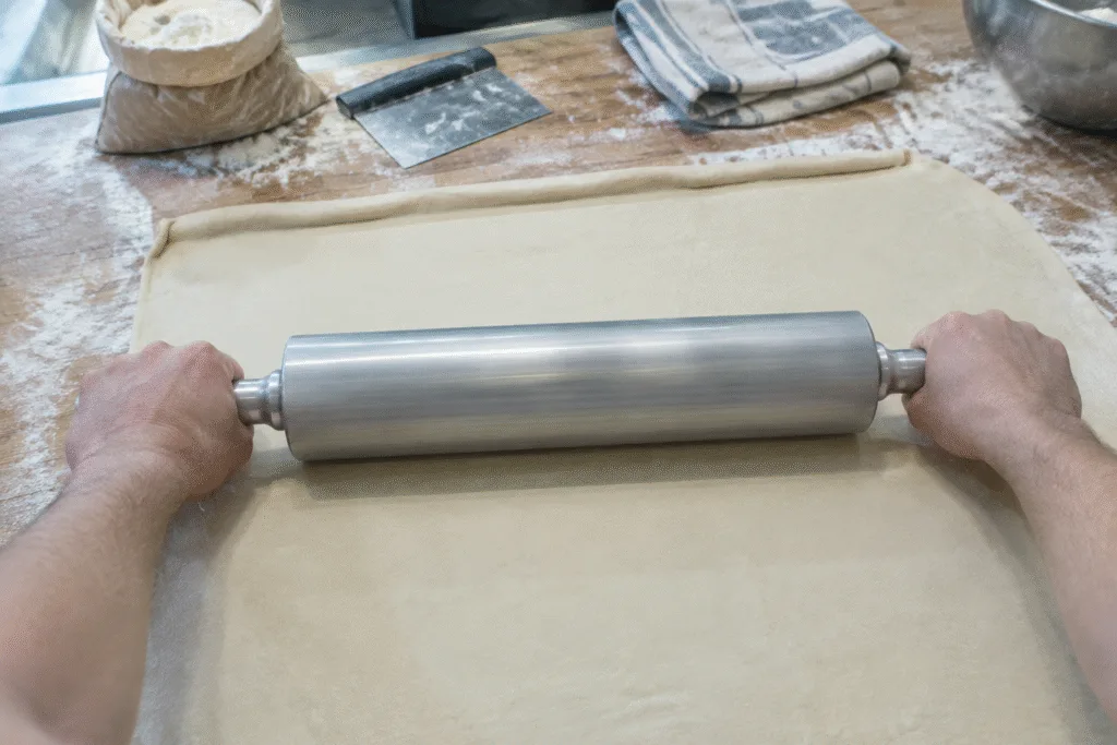 Two hands rolling a large sheet of dough with an aluminum rolling pin on a floured bakery bench